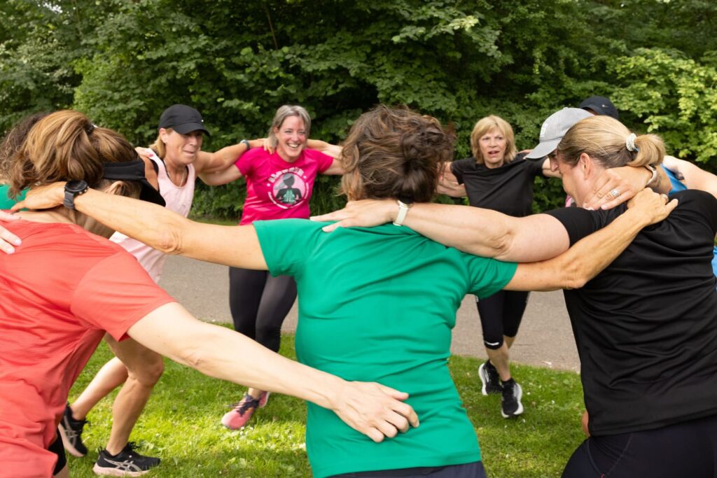 Groep volwassenen in sportkleding vormt een kring op een grasveld tijdens een buitenoefening.
