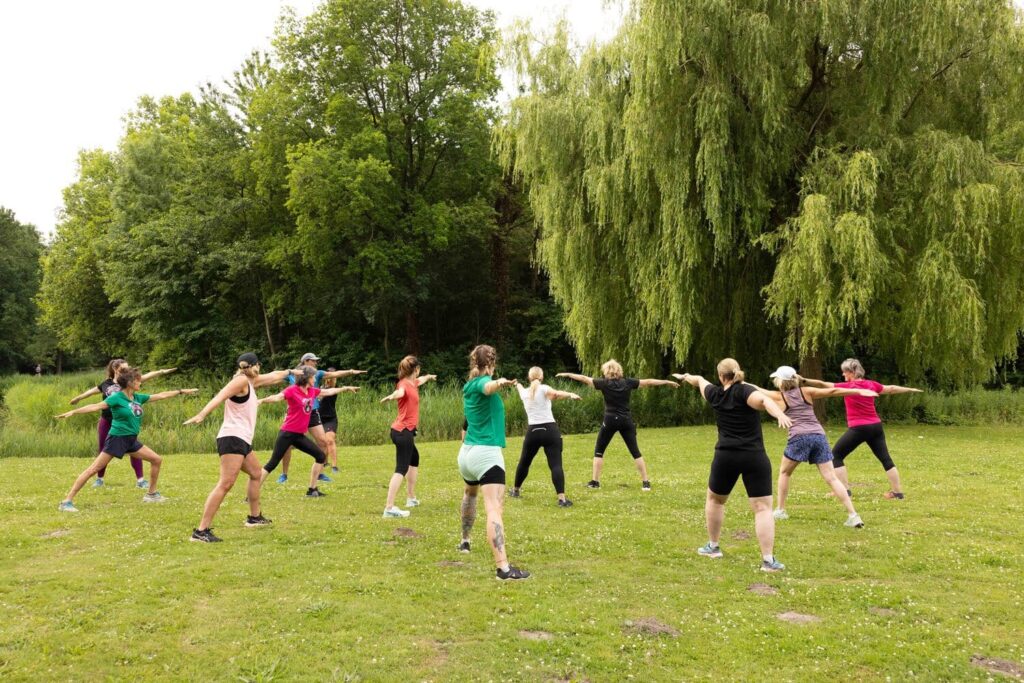 Groep volwassenen doet outdoor fitness in een parkachtige omgeving, omringd door bomen, op een zonnige dag.