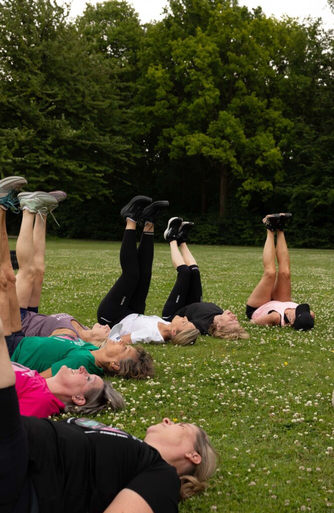 Liggende groep mensen doet stretching oefeningen in park, benen in de lucht gestrekt te midden van groen gras en bomen.